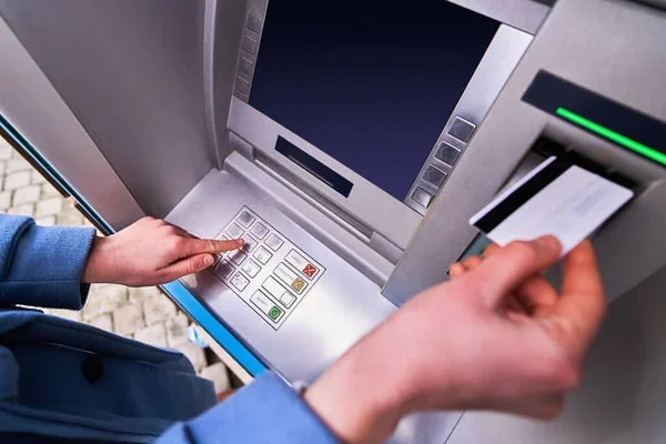 Közelkép egy bankautomatáról, amelynél egy kéz gombokat nyom, a másik pedig bankkártyát tart.